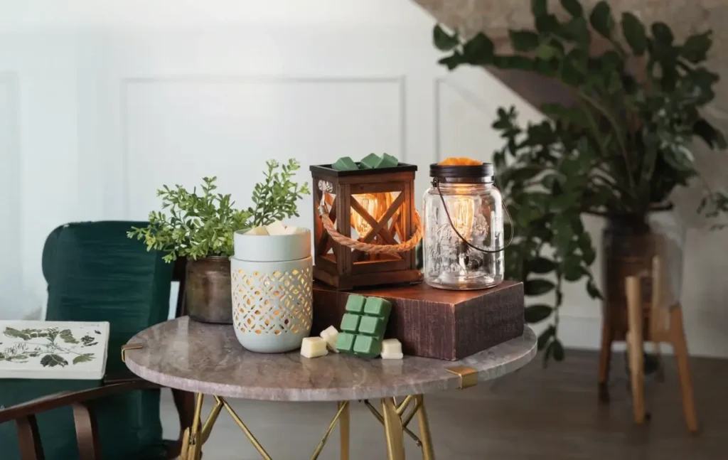 A cozy setup on a marble table featuring various candle warmers, wax melts, and greenery. Includes a decorative wooden lantern, a glass mason jar lamp, and wax cubes in shades of green and cream, creating a warm and inviting ambiance.