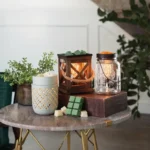 A cozy setup on a marble table featuring various candle warmers, wax melts, and greenery. Includes a decorative wooden lantern, a glass mason jar lamp, and wax cubes in shades of green and cream, creating a warm and inviting ambiance.