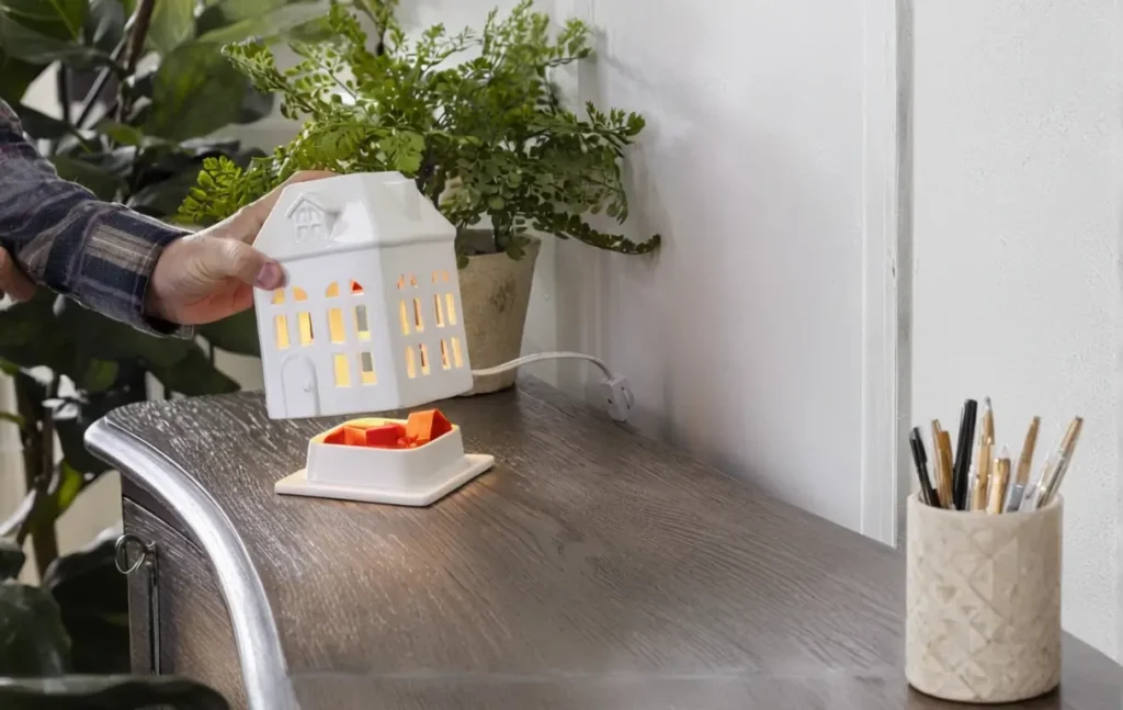 A hand places a small white ceramic house-shaped wax warmer on a wooden tabletop, with bright orange wax cubes melting underneath. The scene is complemented by a pot of green plants in the background and a cup holding pencils and pens nearby.