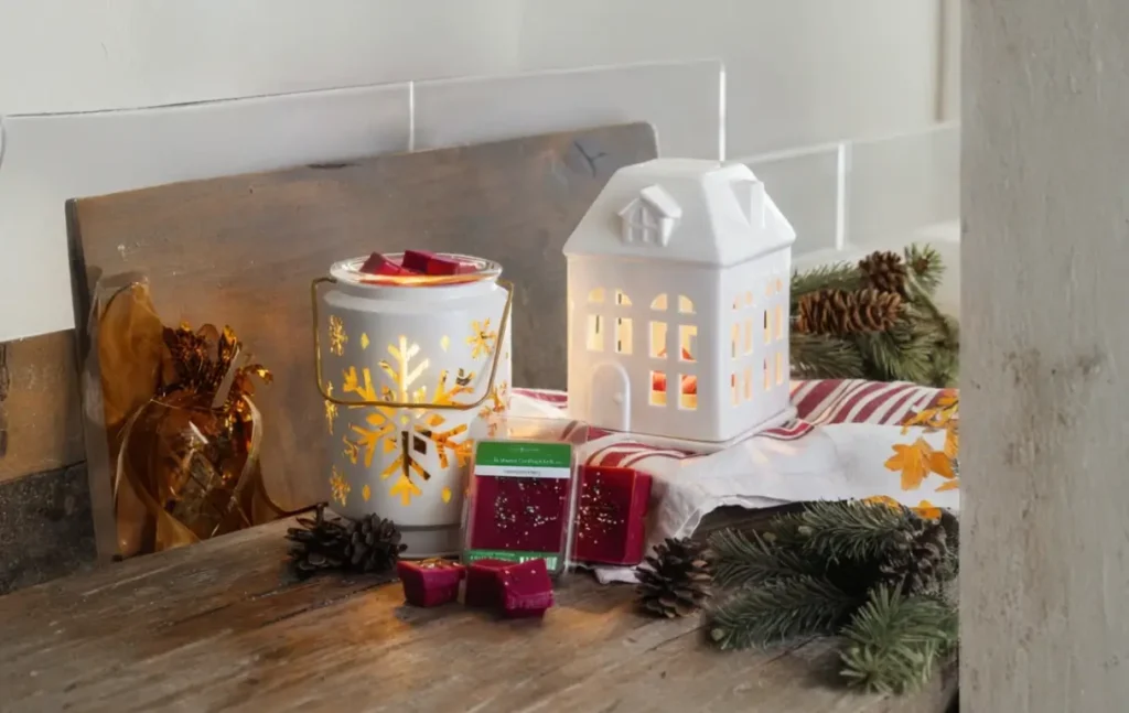 A rustic winter-themed candle warmer setup with a white ceramic house and a snowflake-decorated warmer on a wooden surface. Pine cones, evergreen branches, and red wax melts enhance the cozy, festive atmosphere.